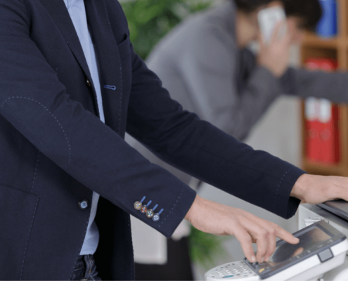 A man pressing buttons on a printer with a coworker on the phone in the background